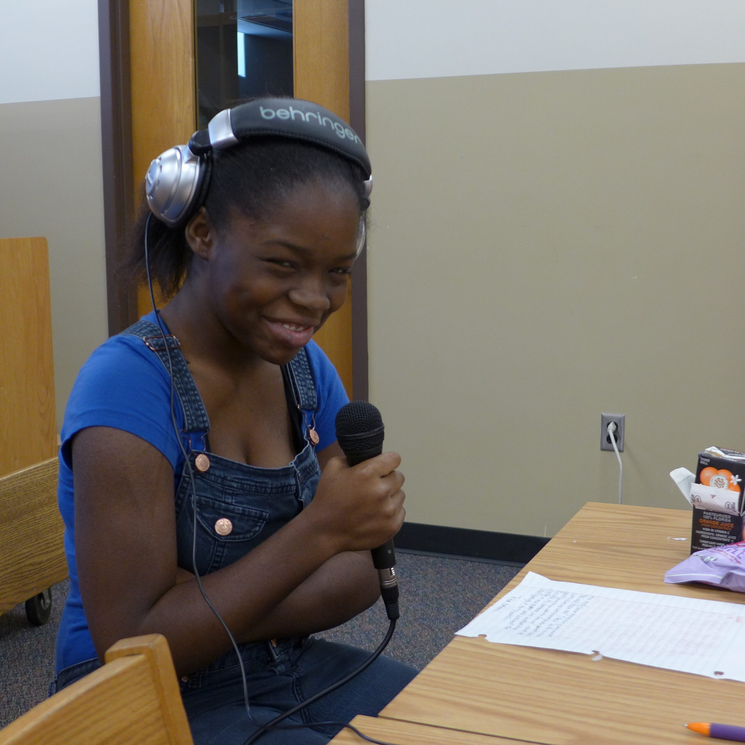 A youth smiling while wearing headphones holding a microphone looking down at a piece of paper on a desk in a classroom