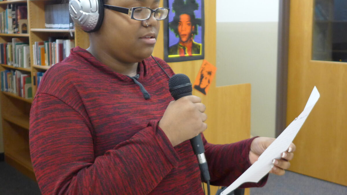 A teen wearing headphones holding a piece of paper and a microphone in a library