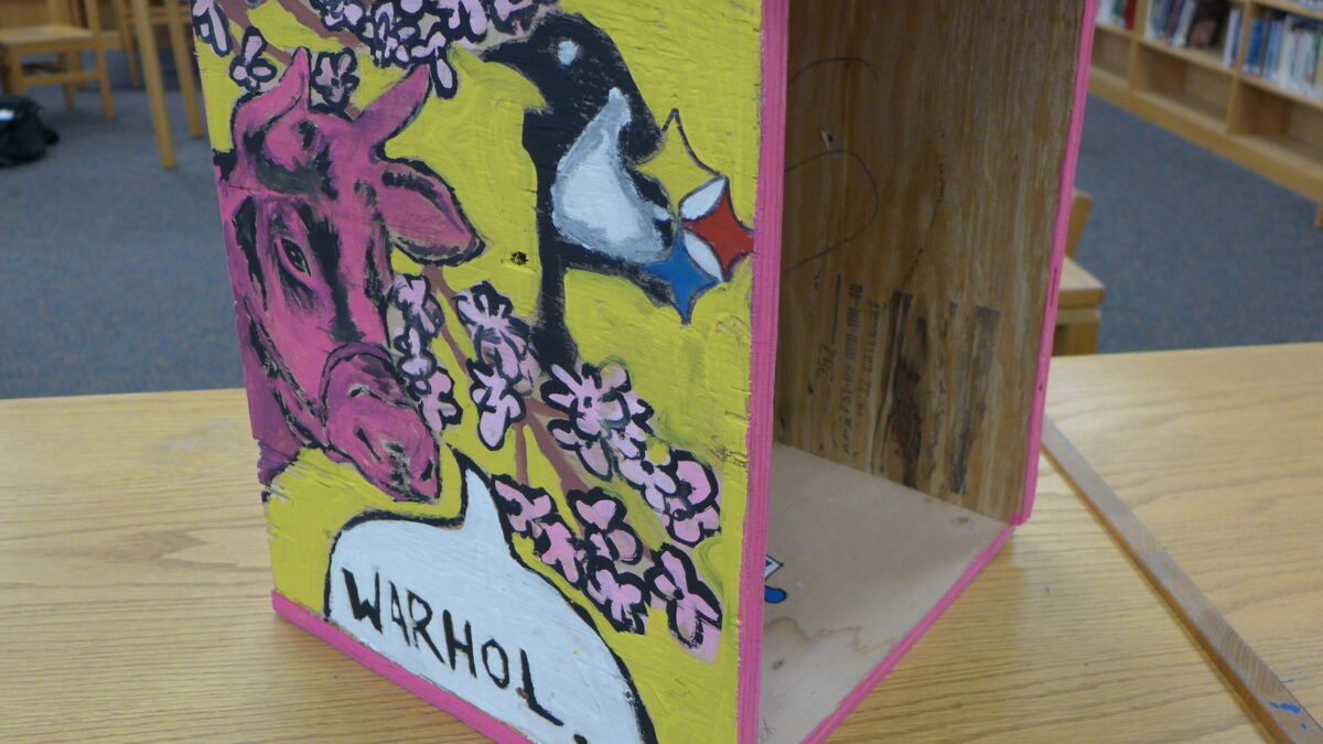 A yellow wooden box with a pink bull, a penguin, and pink flowers painted on in a library