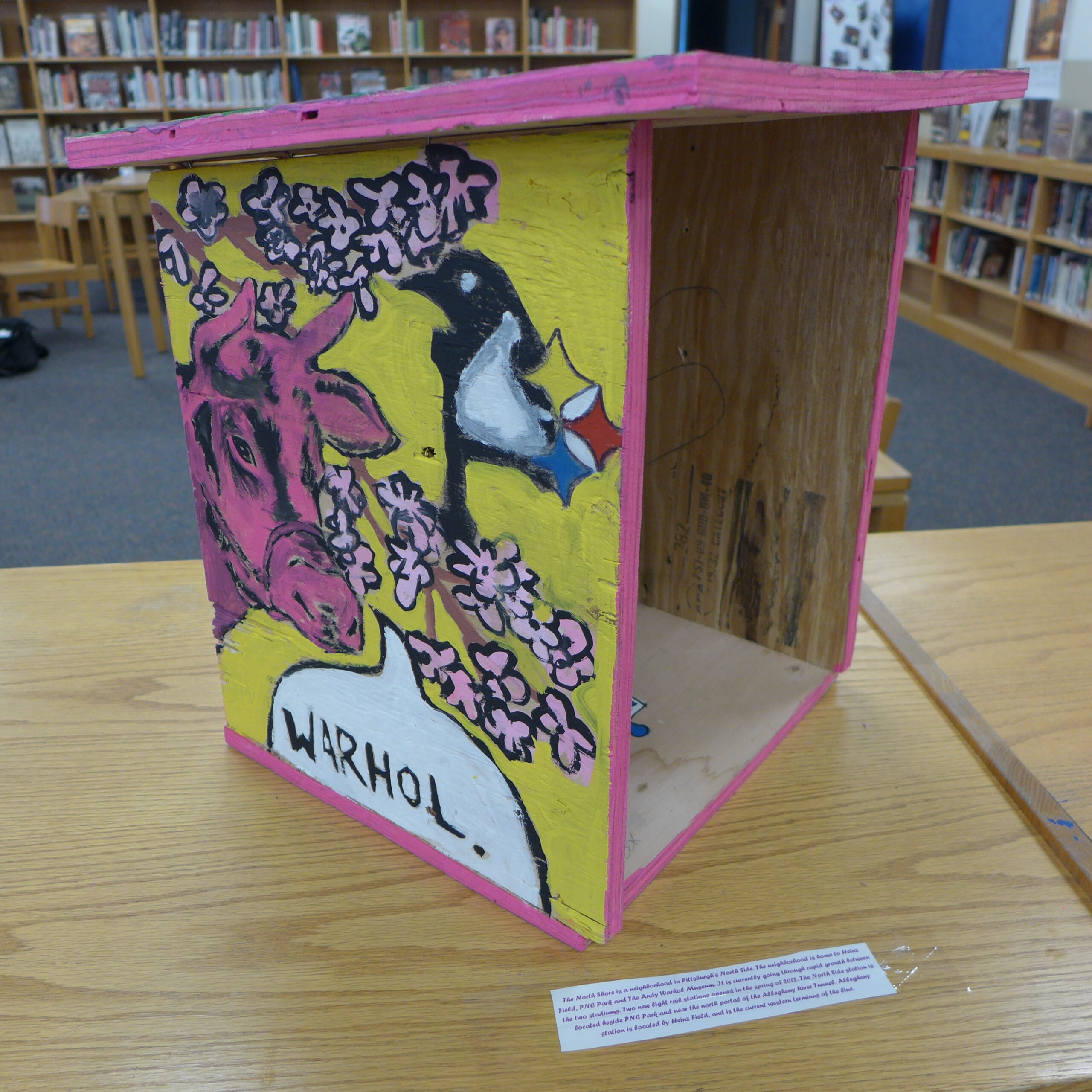 A yellow wooden box with a pink bull, a penguin, and pink flowers painted on in a library