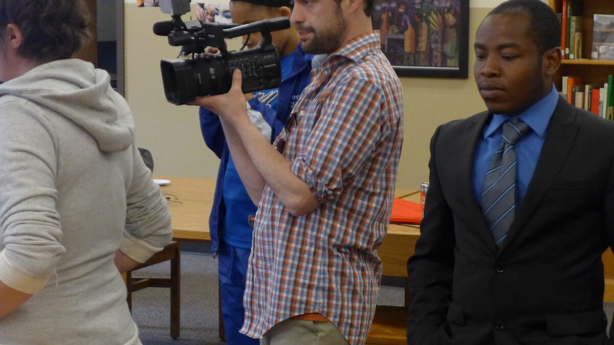 Four adults standing in a library, one holding a video camera
