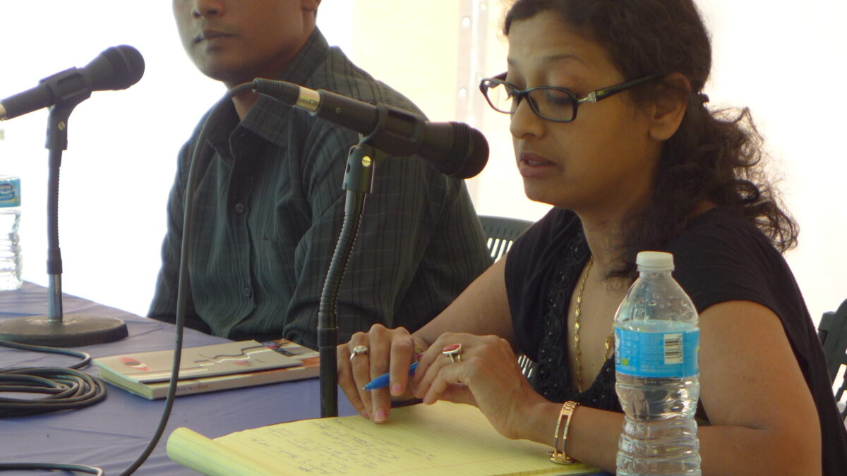 Two adults sitting behind microphones, one looking down at a yellow notepad