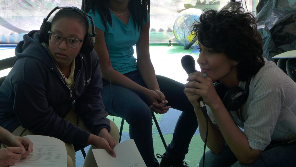 Two youth wearing headphones sitting across from an adult wearing headphones around their neck and holding a microphone