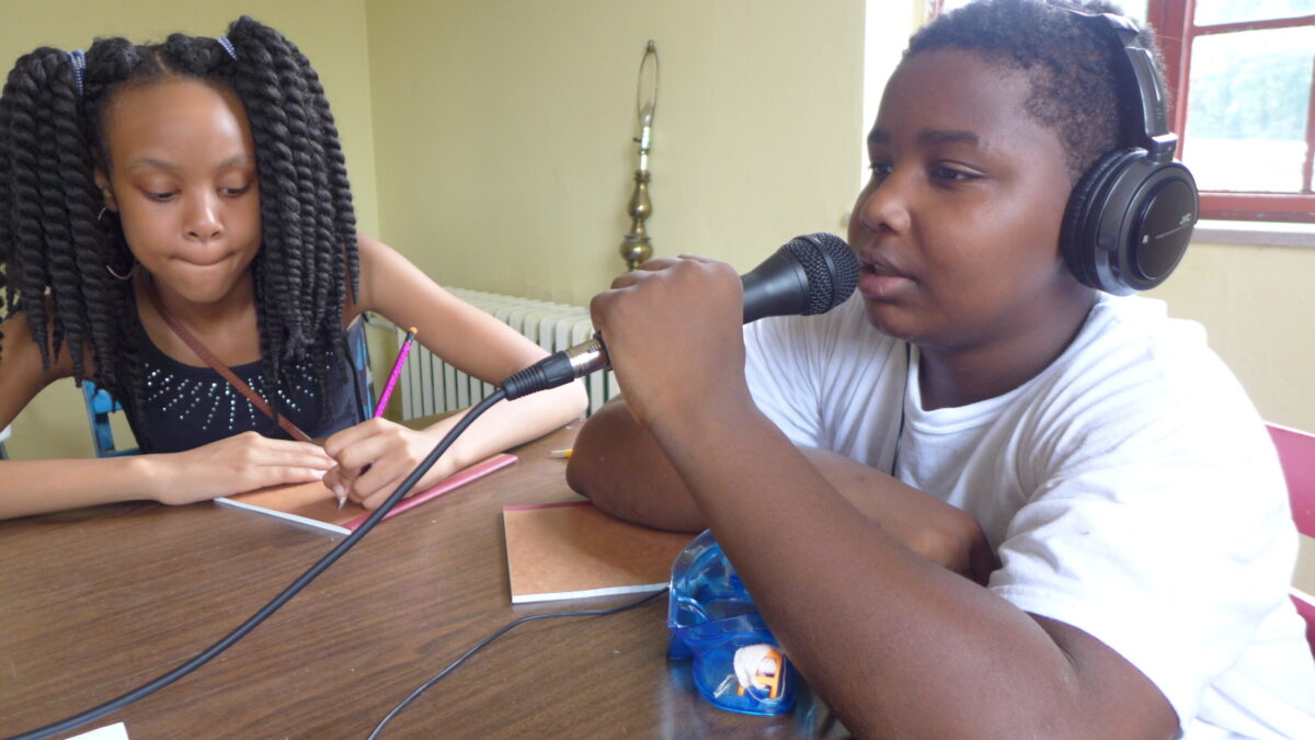 A youth writing on a notepad and a youth wearing headphones holding and speaking into a microphone