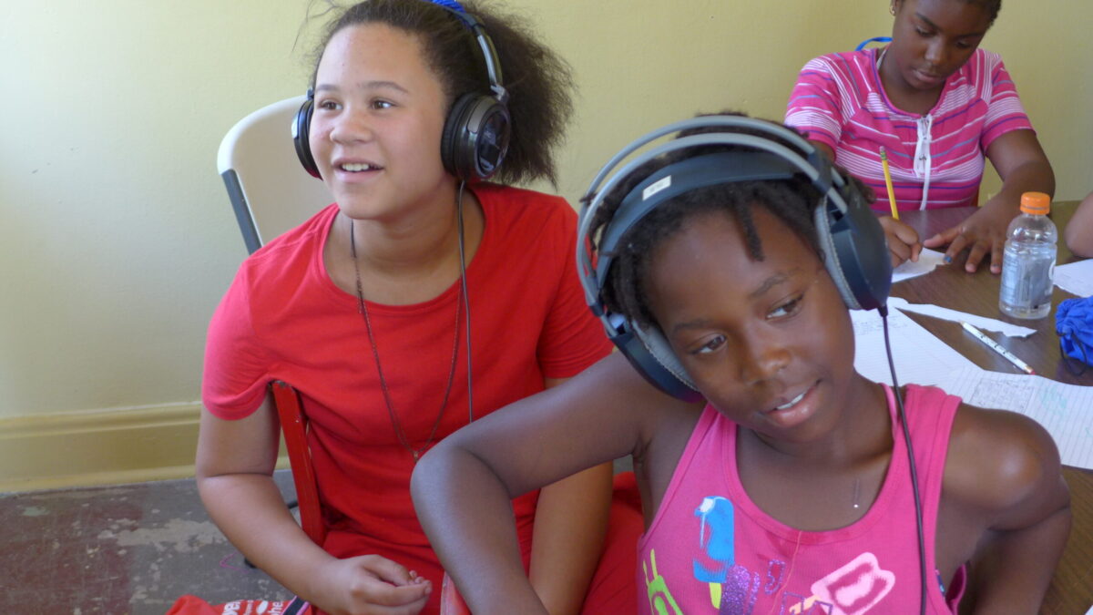 Two youth wearing headphones and sitting and a youth writing on a piece of paper on a brown table