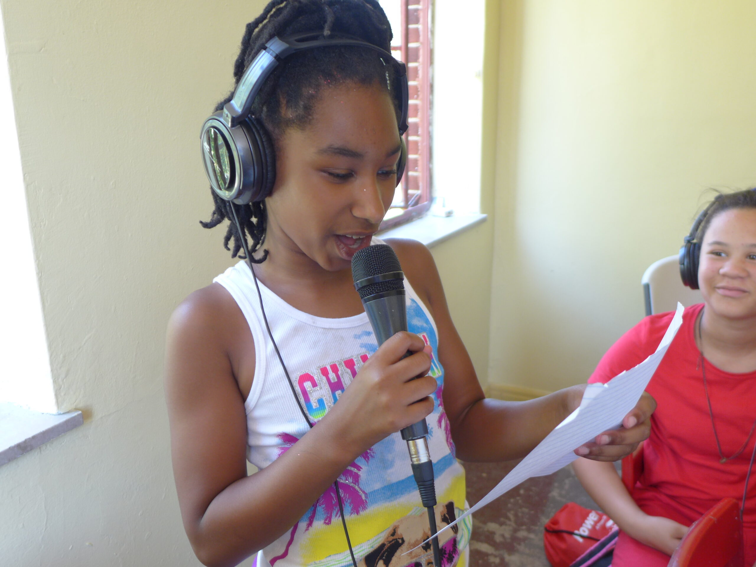 A youth wearing headphones looking at another youth wearing headphones holding a piece of paper and speaking into a microphone