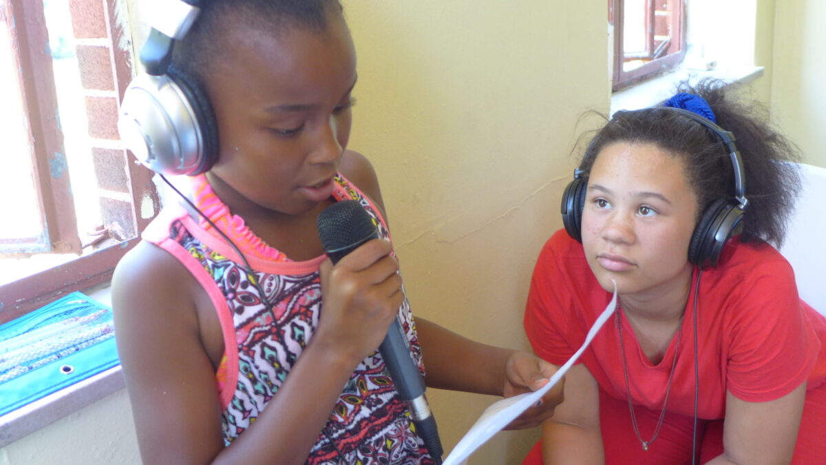 A youth wearing headphones looking at another youth wearing headphones holding a piece of paper and speaking into a microphone