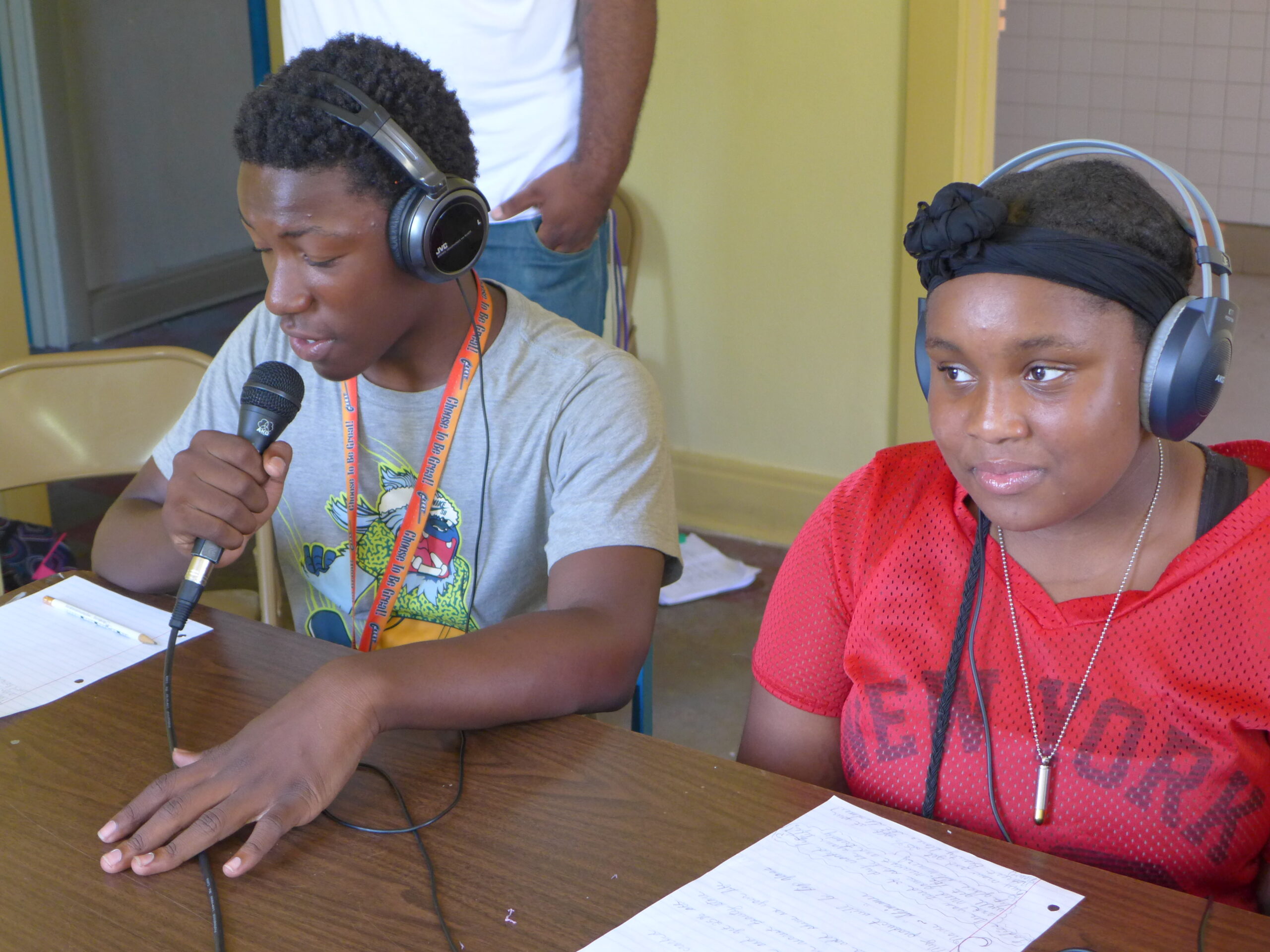 Two youth wearing headphones sitting at a brown table, one holding and speaking into a microphone