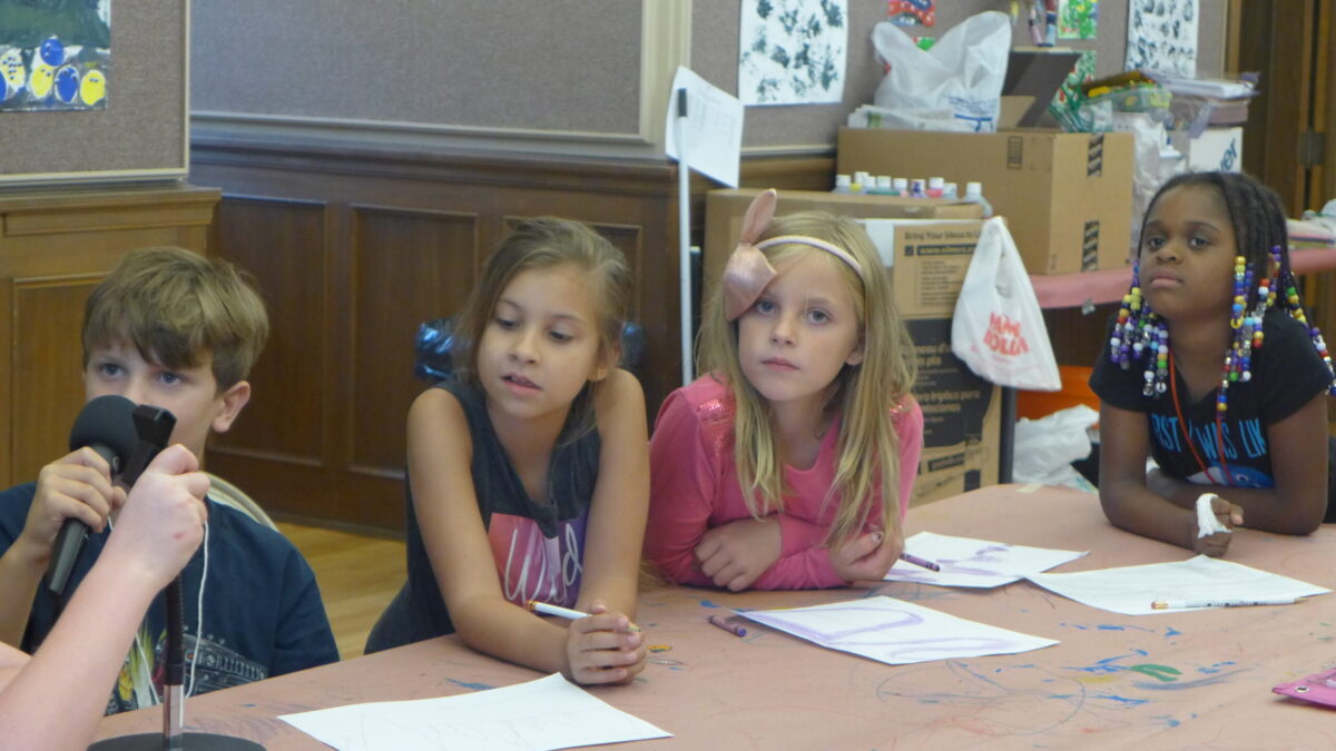 Four youth sitting behind pieces of paper on a table, one holding a microphone