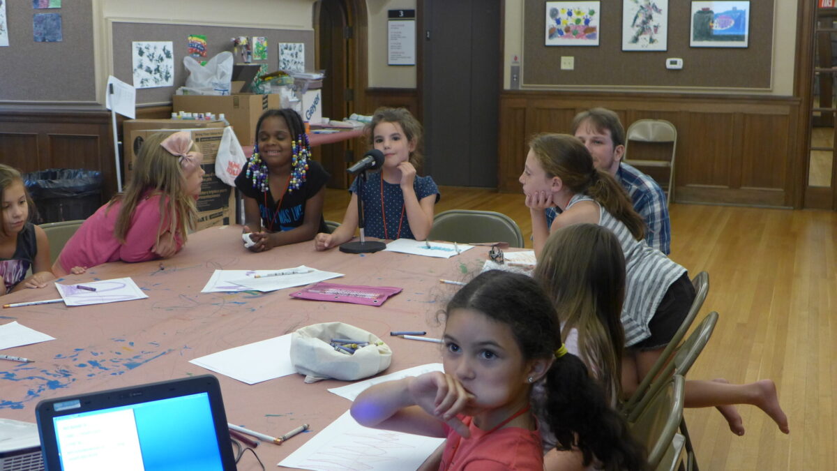 Eight youth sitting around pieces of paper on tables