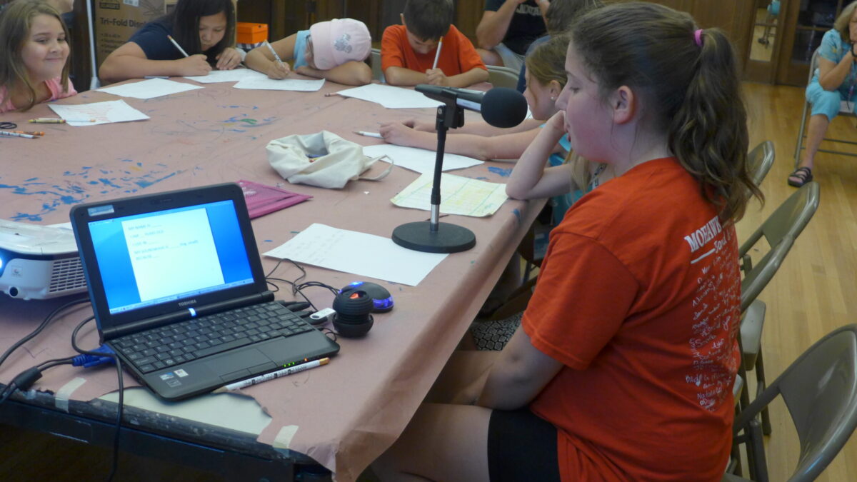 A youth speaking into a microphone as five youth listen while writing on pieces of paper