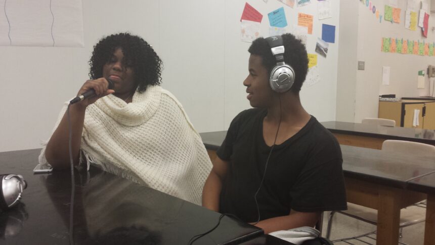 A teen wearing headphones looking at another teen speaking into a microphone in a classroom