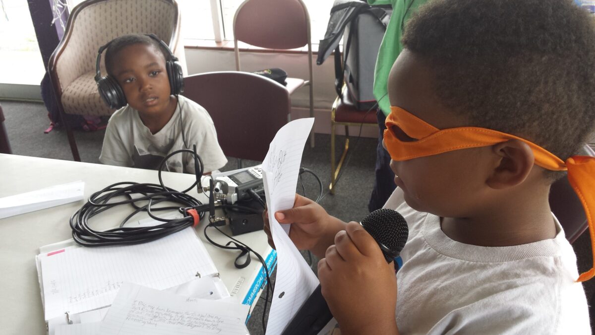 20150706_103235 A youth wearing an orange eye mask reading a piece of paper and holding a microphone and another youth sitting nearby behind a long white table