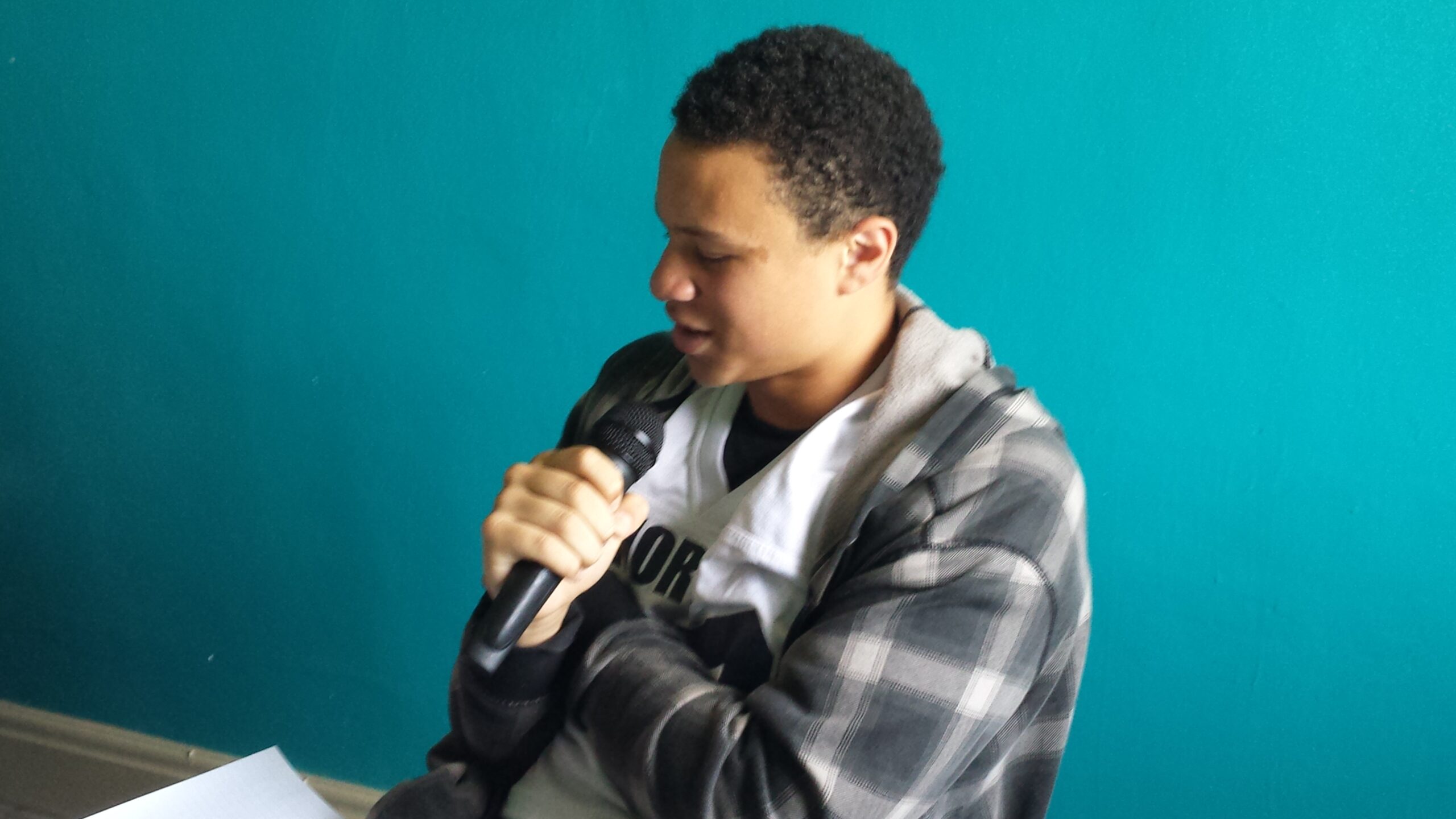 A youth holding and speaking into a microphone and holding a piece of paper in a blue room