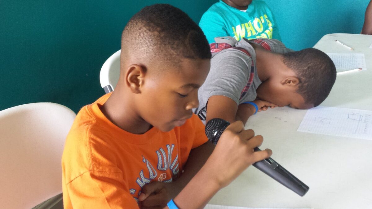 A youth holding a microphone looking down and a youth face down on a white table in a blue room