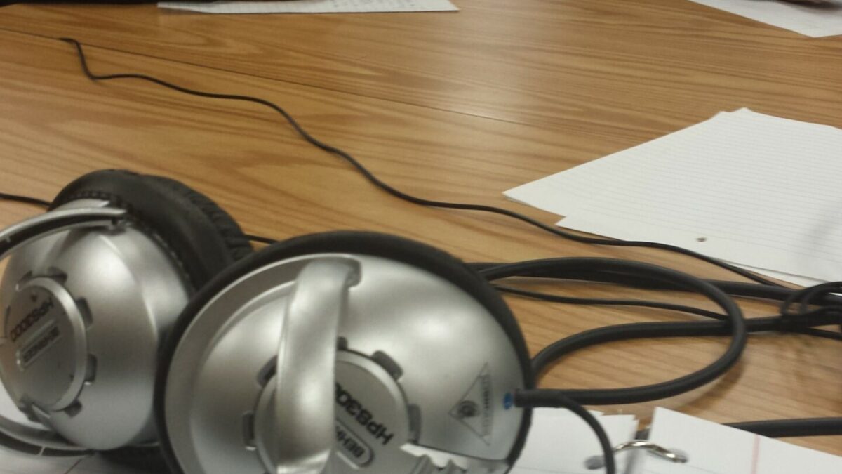 A closeup of a pair of headphones with two youth, one wearing headphones, writing on pieces of paper in the background in a library