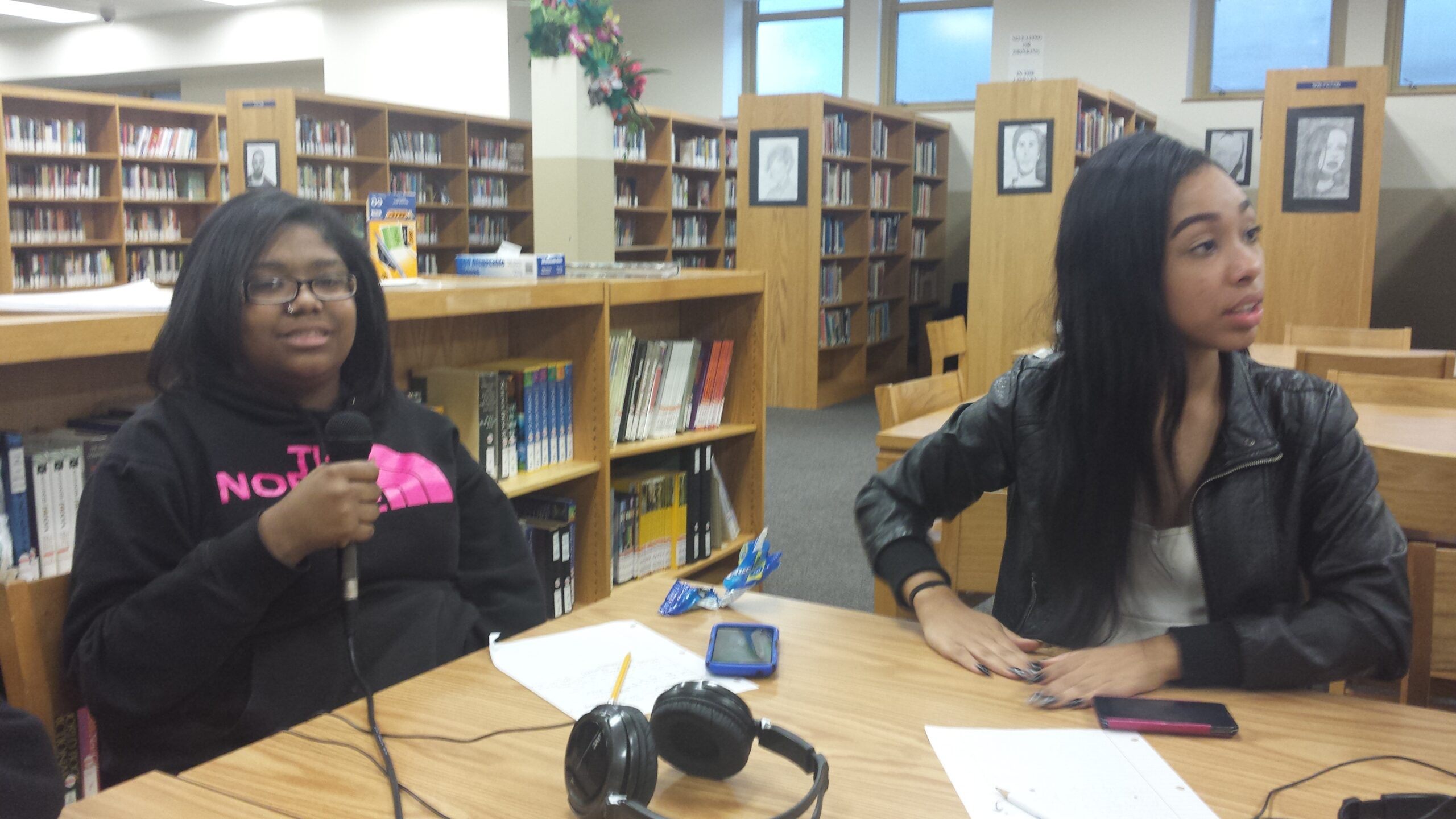 Two teens, one holding a microphone, sitting at a table in a library