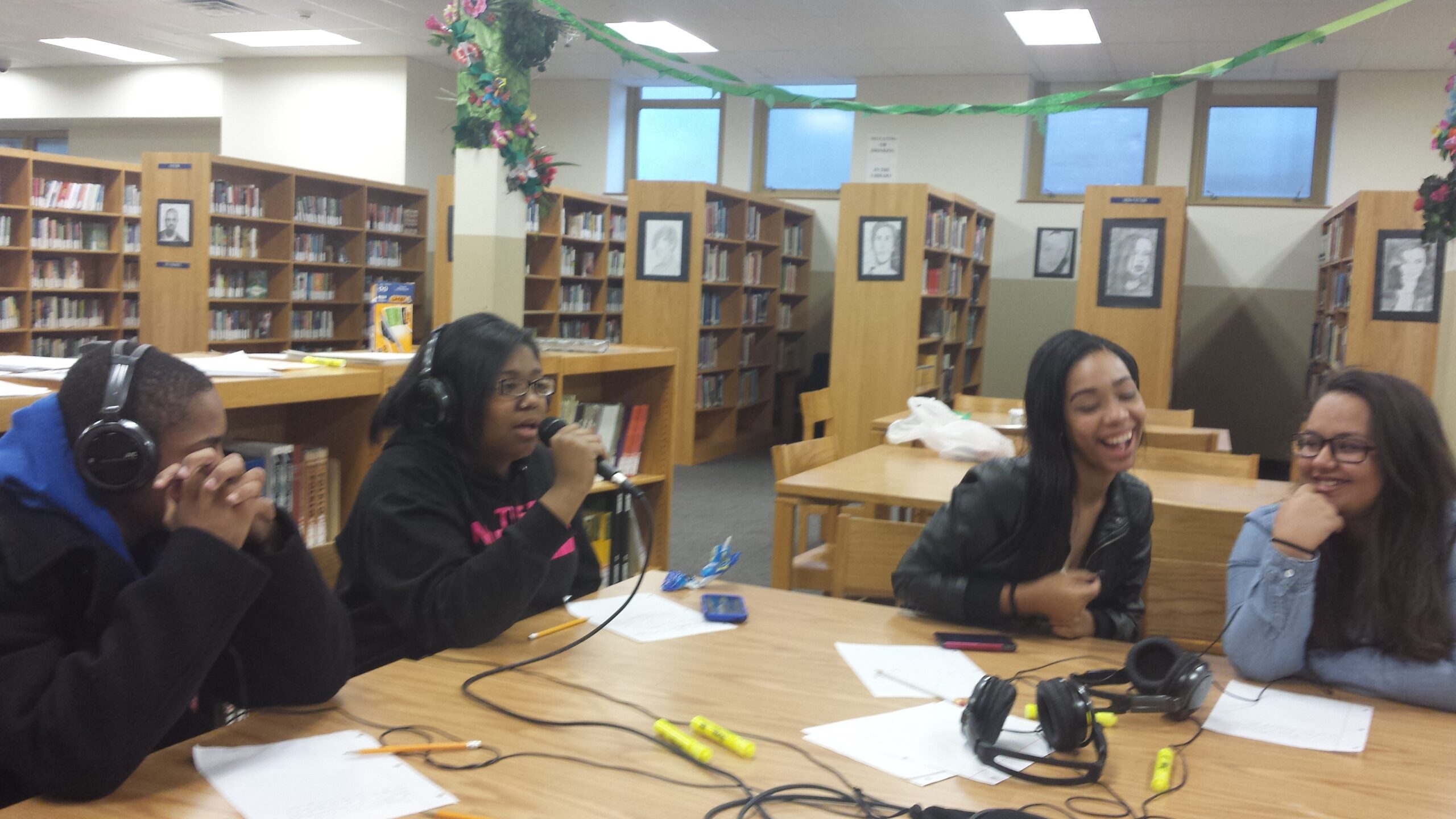 Four teens, two wearing headphones and one holding a microphone, sitting at a table in a library