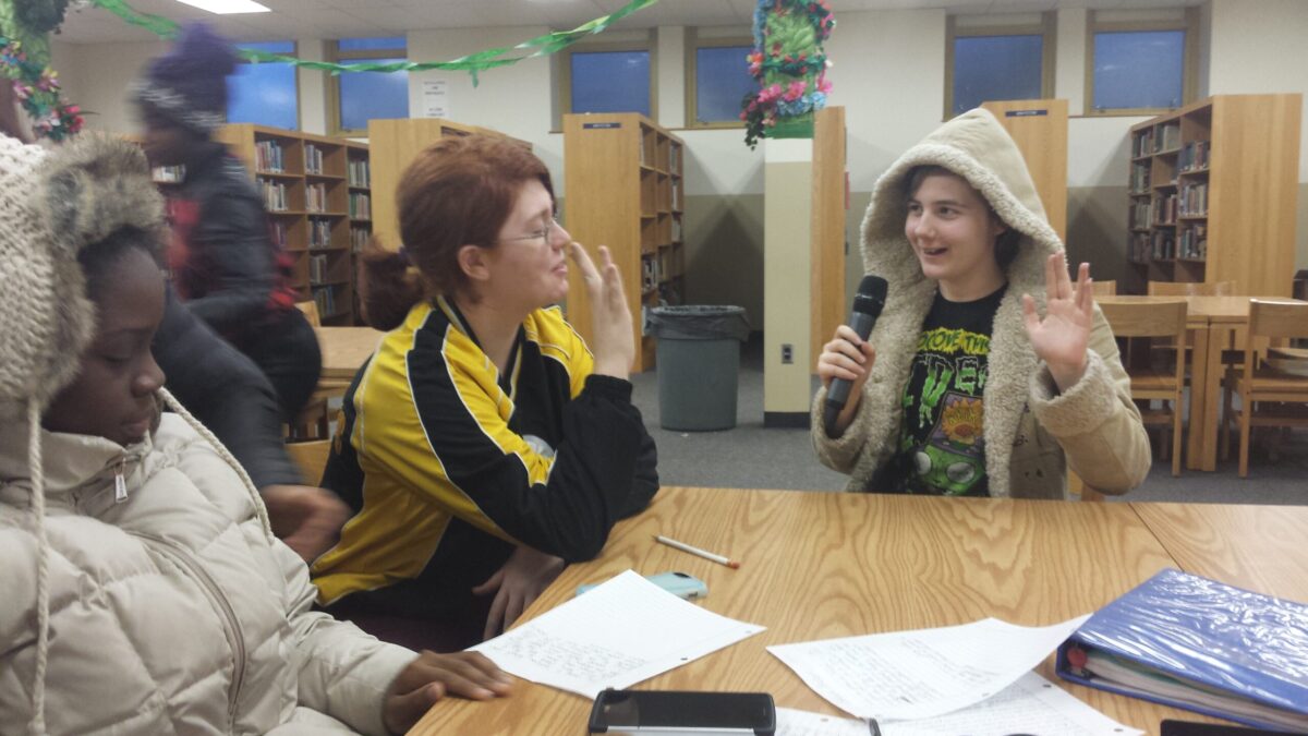 20151201_162955 Three teens sitting at a wooden table in a library, one teen holding and speaking into a microphone