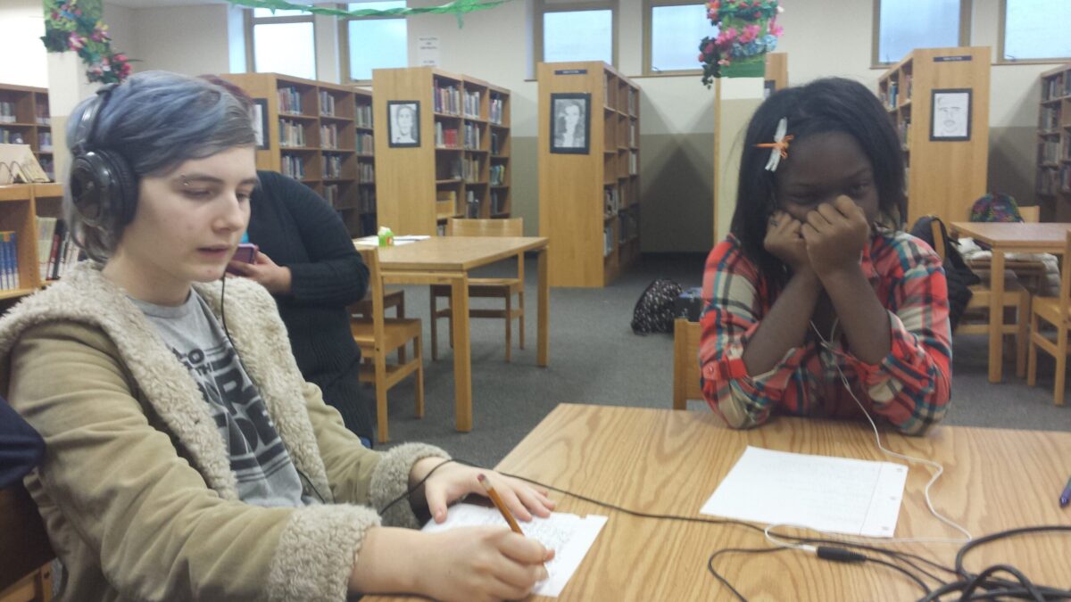 20151208_160534 Two teens, one wearing headphones, sitting behind pieces of paper on a wooden table in a library