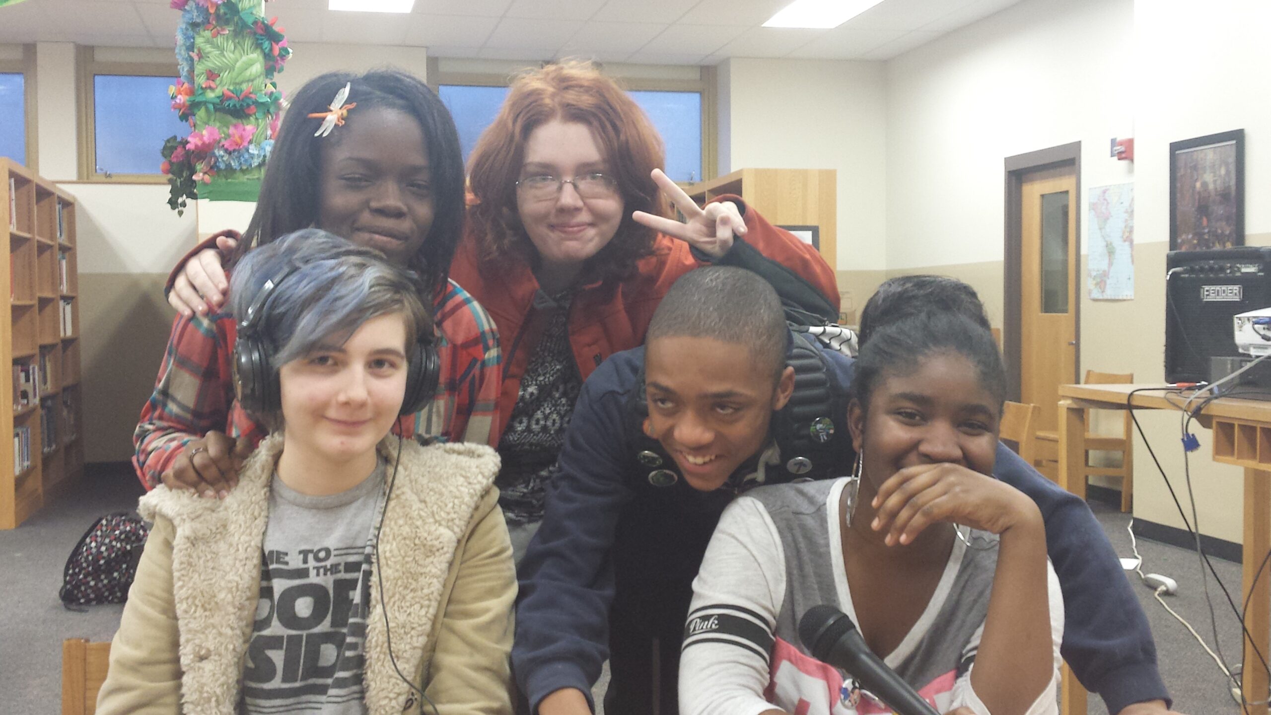 Five teens smiling and posing behind a wooden table in a library