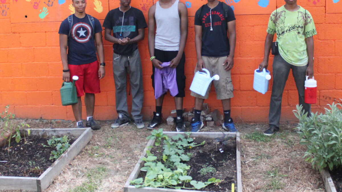 Group-Image Five teens holding watering cans standing in front of an orange wall covered in multicolored handprints.