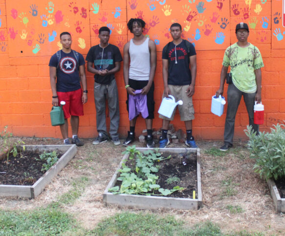 Five teens holding watering cans standing in front of an orange wall covered in multicolored handprints.