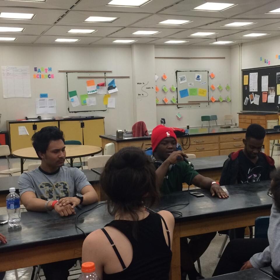 Four teens sitting around a desk in a classroom, one holding and speaking into a microphone