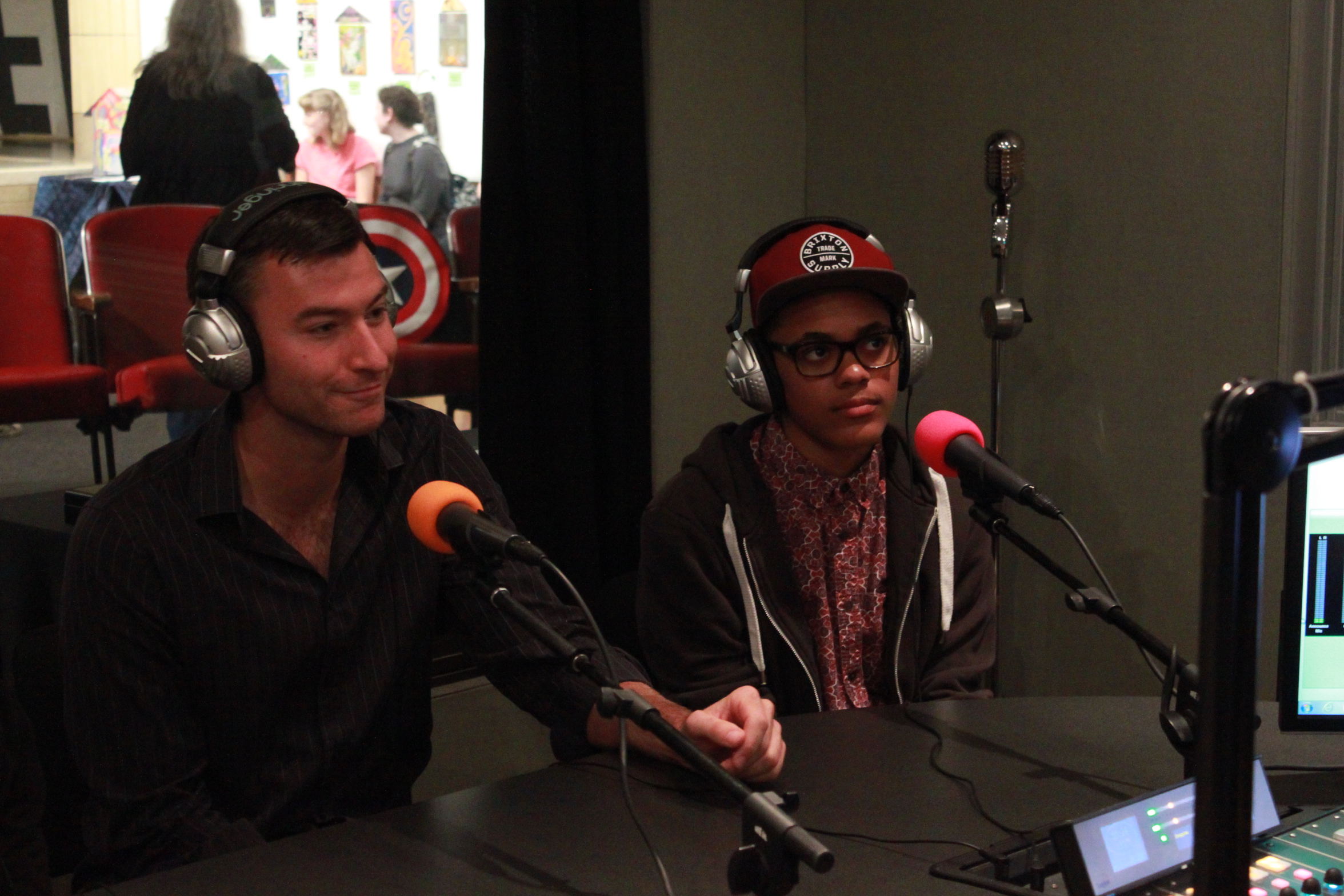 An adult and a teen wearing headphones sitting behind microphones in a radio studio