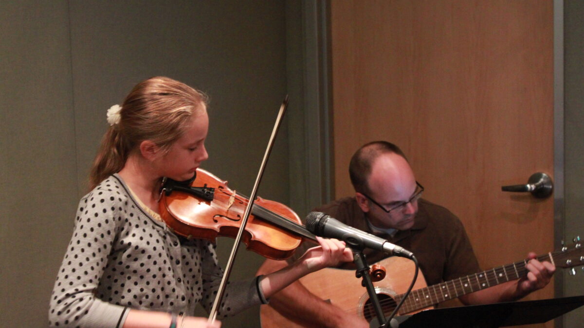 A youth playing a violin and an adult playing an acoustic guitar in a radio studio