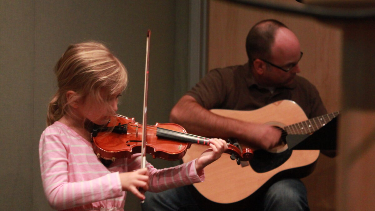 A youth playing a violin and an adult playing an acoustic guitar in a radio studio