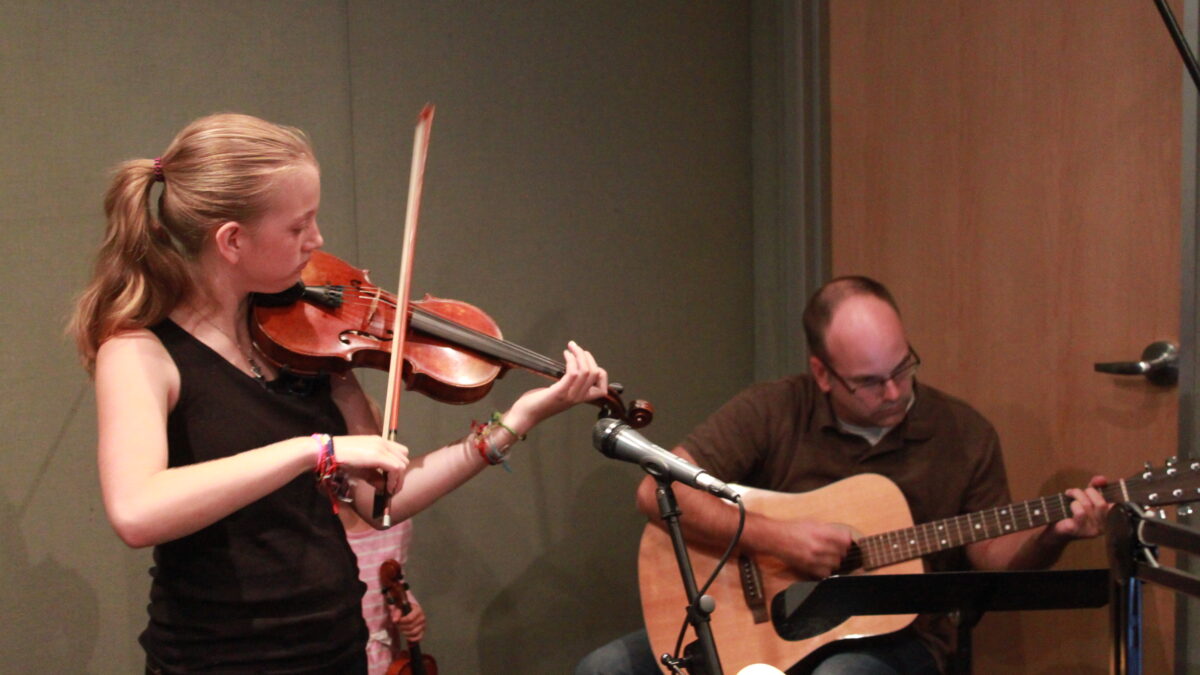A youth playing violin and an adult playing acoustic guitar in a radio studio