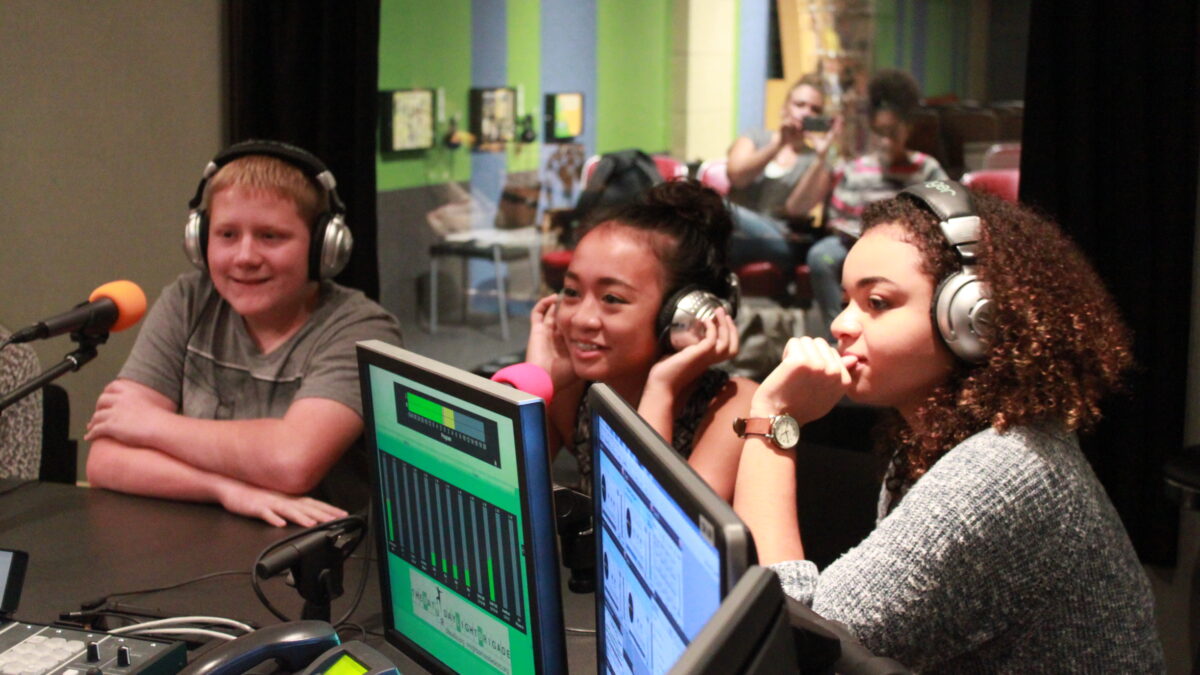 Three teens wearing headphones speaking into microphones in a radio studio