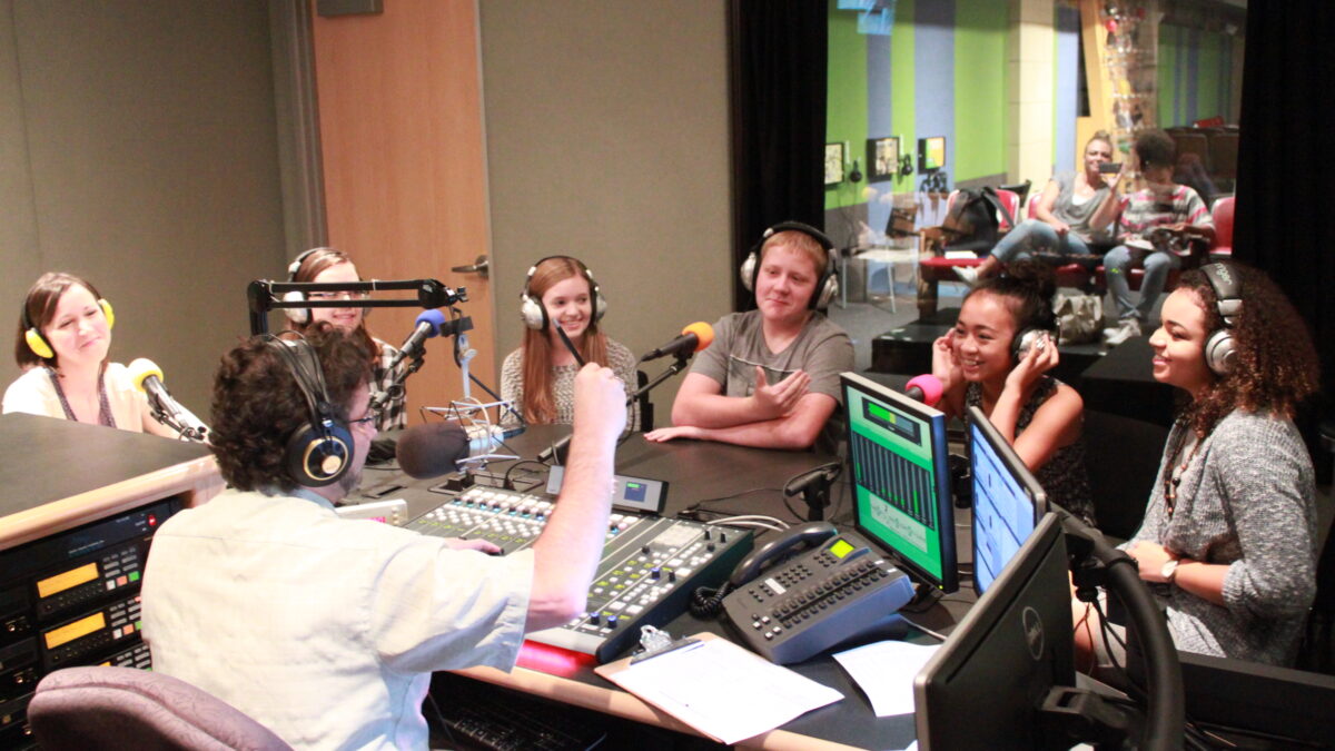 Six teens wearing headphones speaking into microphones being interviewed by an adult wearing headphones sitting behind a microphone and a mixing board in a radio studio