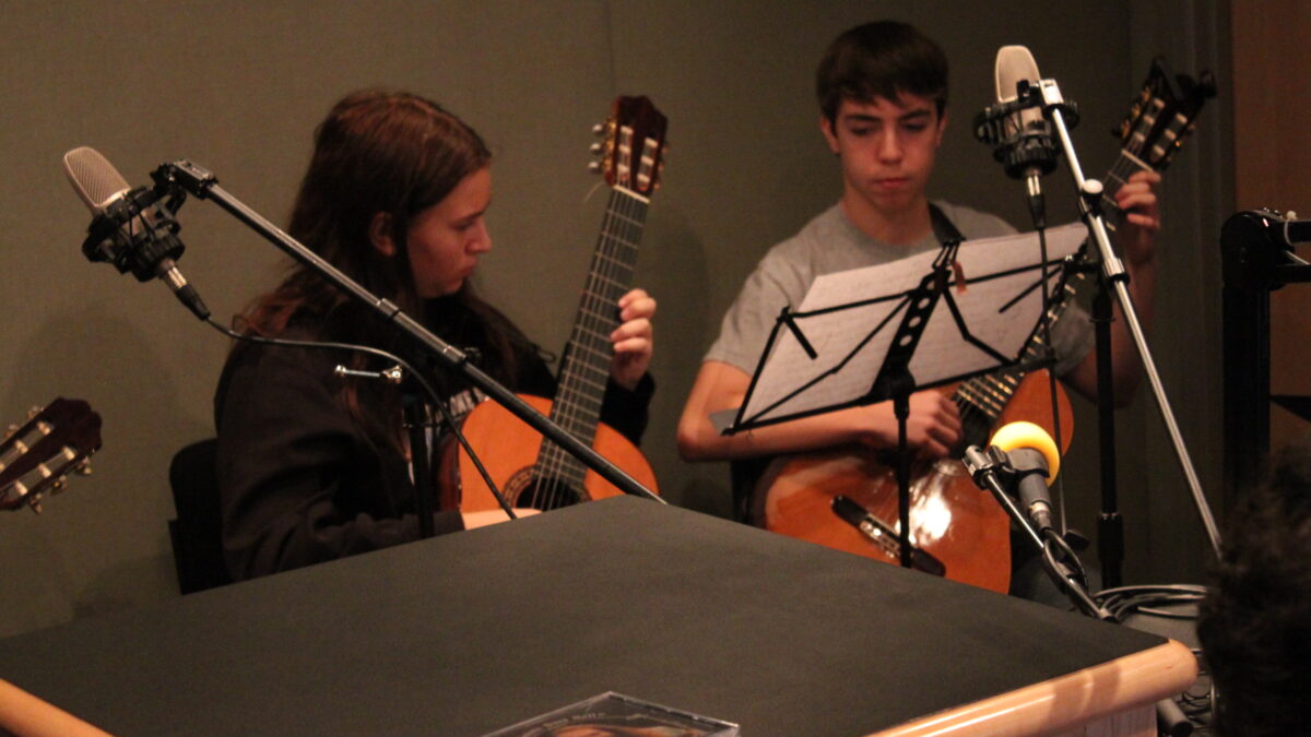 Two youth playing acoustic guitars looking at sheet music on music stands in a radio studio
