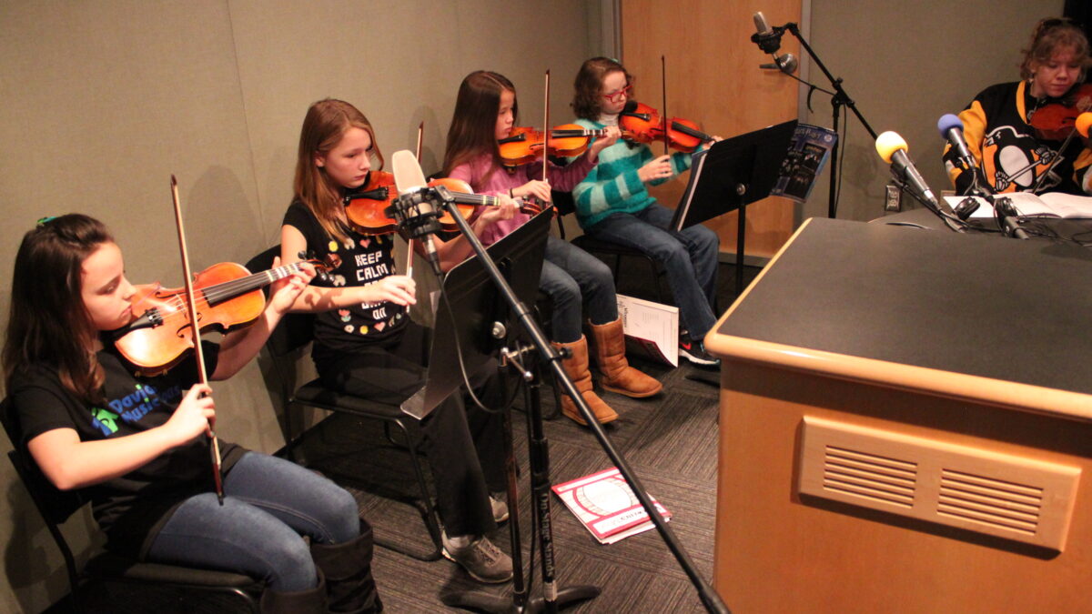IMG_0444 Four youth and an adult playing violins looking at sheet music on music stands in a radio studio