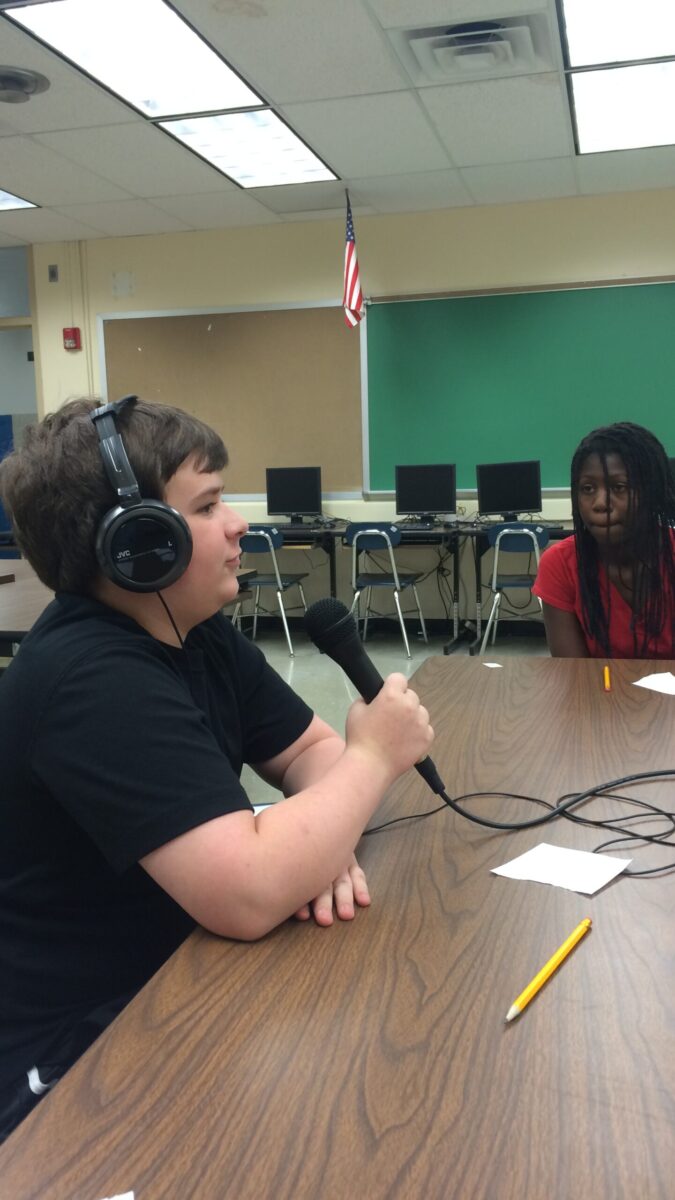 Two youth sitting around a brown table in a classroom, one wearing headphones and holding and speaking into a microphone