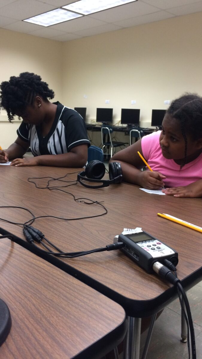 Two youth writing on pieces of paper on a brown table in a classroom