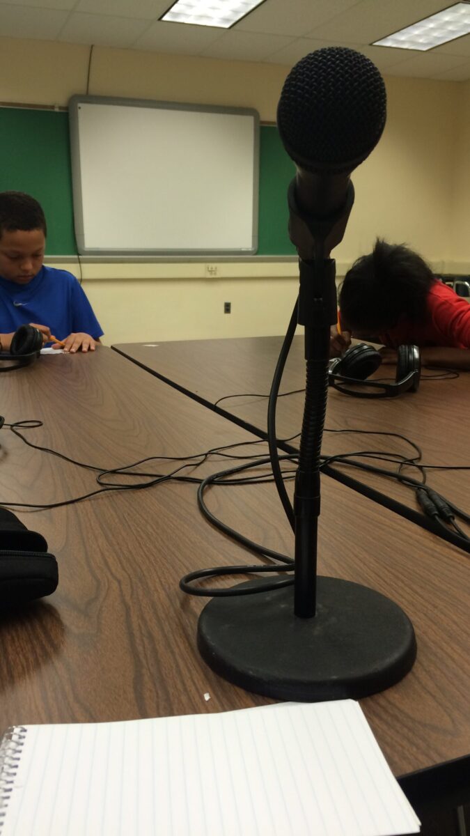 A closeup of a microphone and two youth writing on pieces of paper on a brown table in the background
