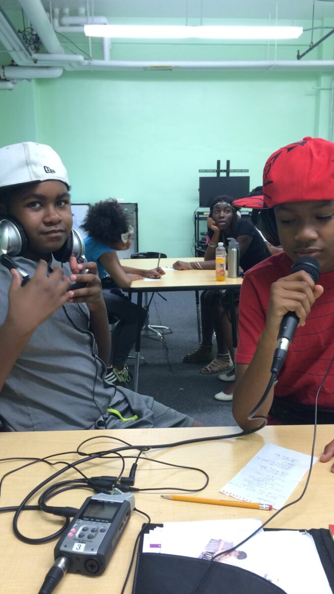Two youth wearing headphones and baseball caps sideways sitting at a table, one youth speaking into a microphone connected to a portable recorder