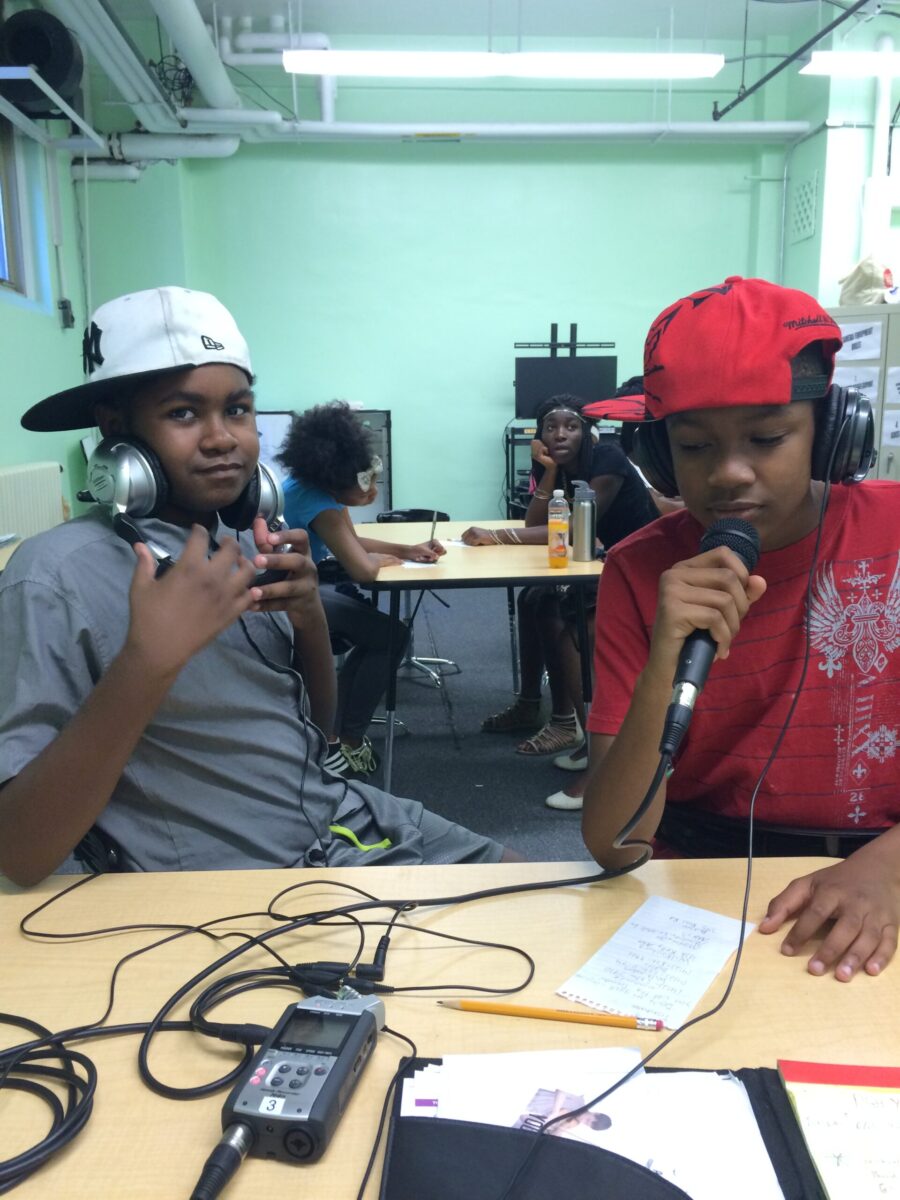 Two youth wearing headphones and baseball caps sideways sitting at a table, one youth speaking into a microphone connected to a portable recorder