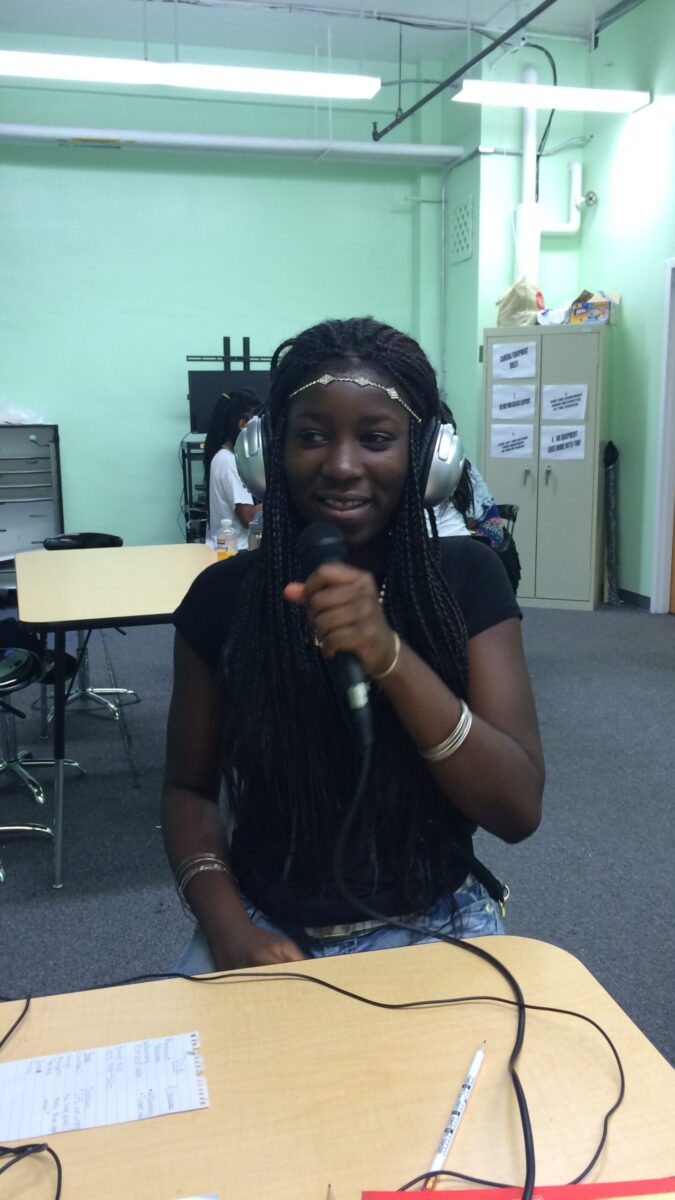 A teen wearing headphones holding and speaking into a microphone while sitting at a classroom desk