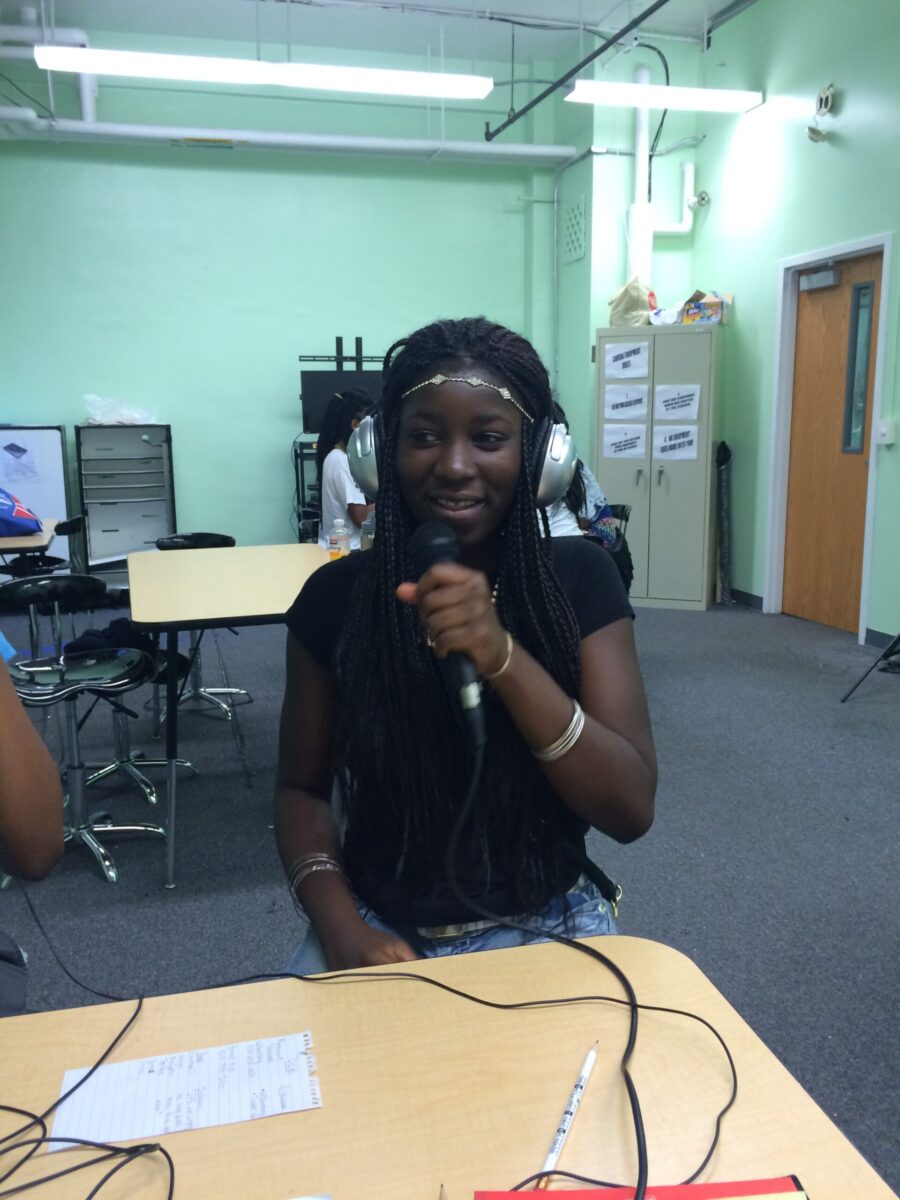 A teen wearing headphones holding and speaking into a microphone while sitting at a classroom desk