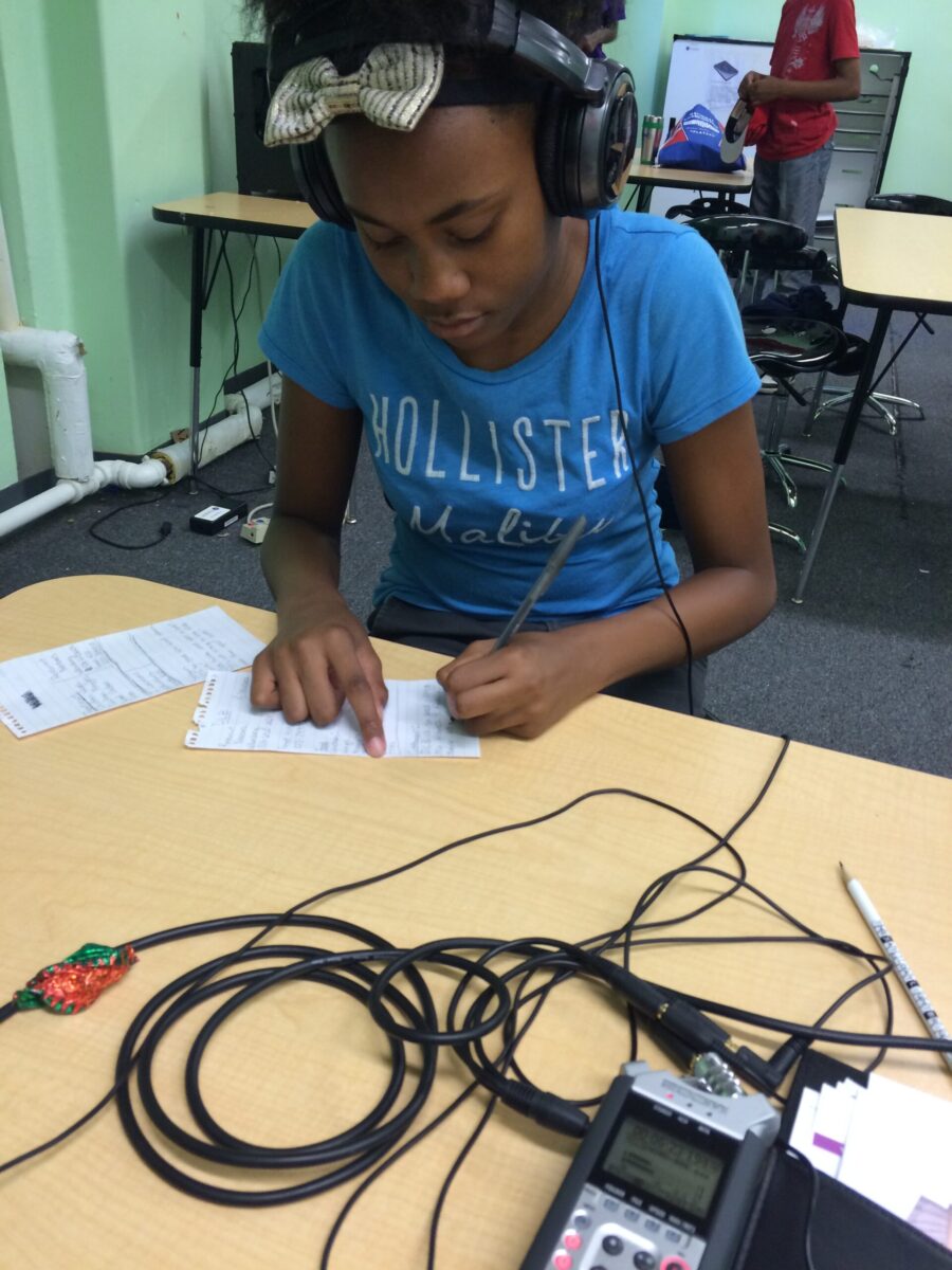 A teen wearing headphones writing on a piece of paper on a classroom desk