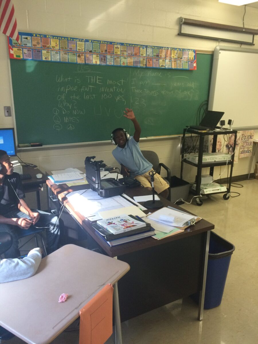 A youth wearing headphones sitting next to a portable recorder raising their hand in front of a chalkboard