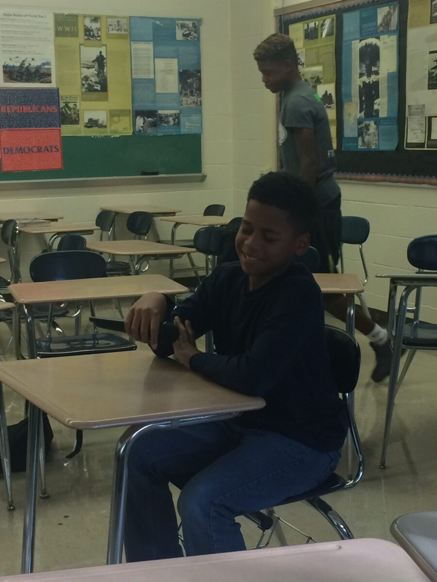 A youth sitting at a classroom desk holding a microphone and smiling