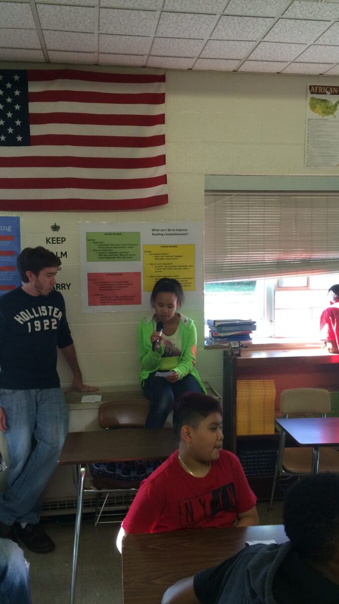 Three youth, one holding a microphone, sitting in a classroom as an adult watches