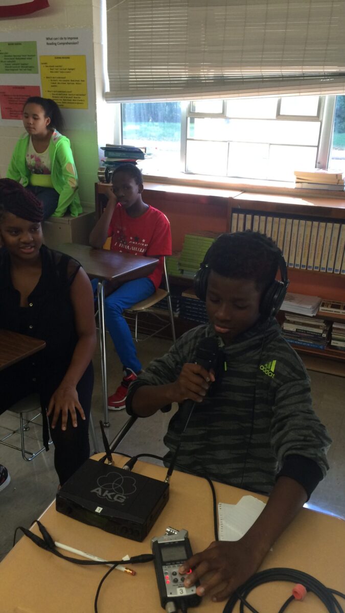 Four youth sitting in a classroom, one wearing headphones speaking into a microphone and interacting with a portable recorder