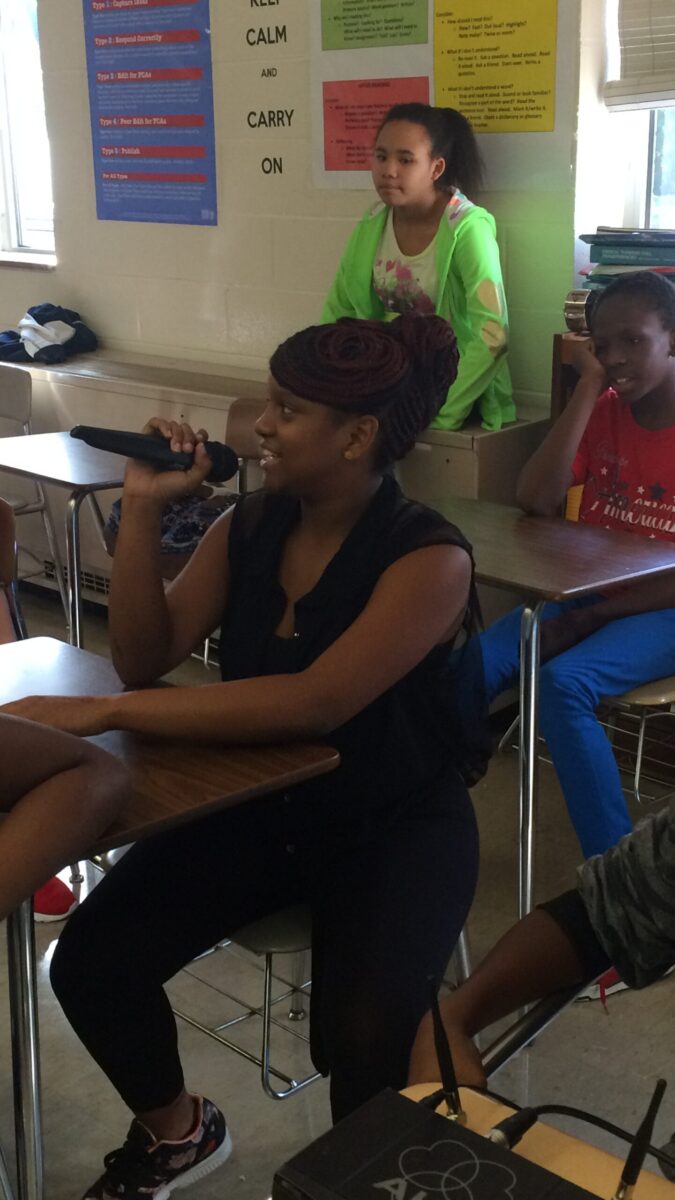 Three youth sitting in a classroom, one holding and speaking into a microphone
