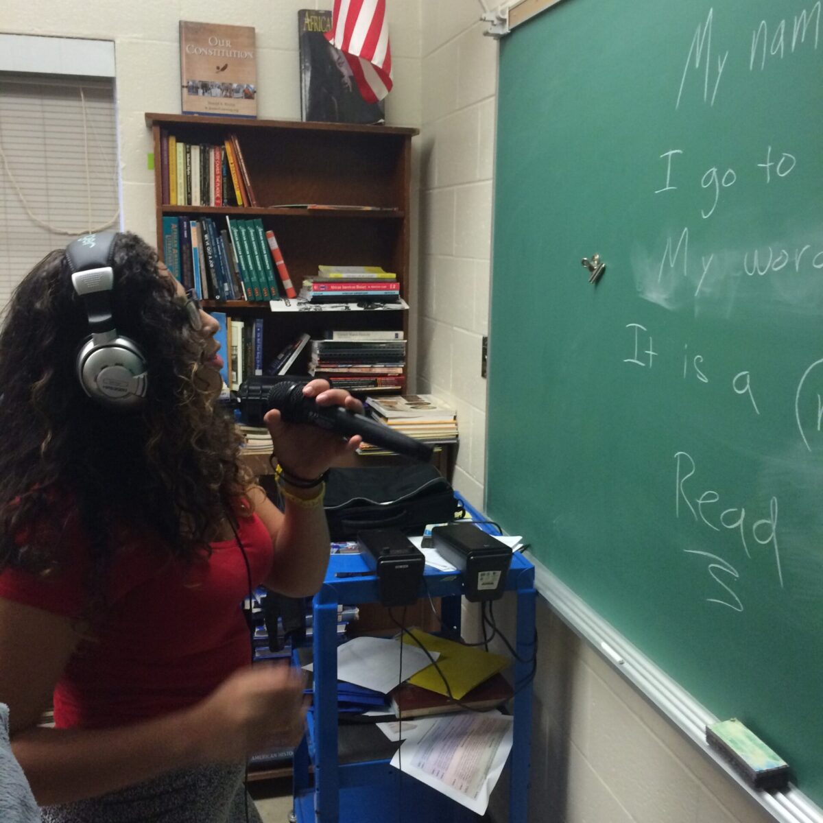A youth wearing headphones holding a microphone and looking at a chalkboard in a classroom
